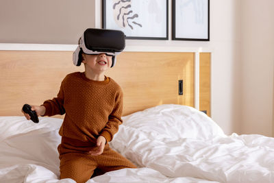 Little adorable boy sitting on bed at home with vr headset and playing interactive video game