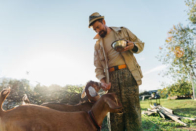 A man farmer in hat feeds nubian goats oats from a bowl on his free-range farm