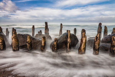 Wooden posts and rocks in sea against cloudy sky