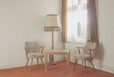 Chairs with table and electric lamp arranged by window at home