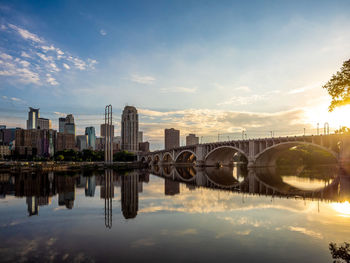 Bridge over river by buildings against sky during sunset