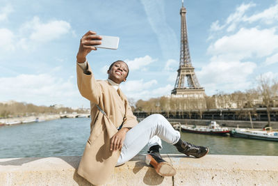 France, paris, woman sitting on bridge over the river seine with the eiffel tower in the background taking a selfie