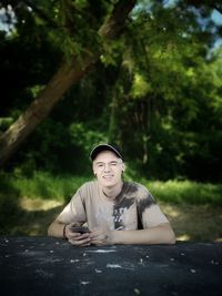 Portrait of young man using mobile phone while sitting at table against trees in park