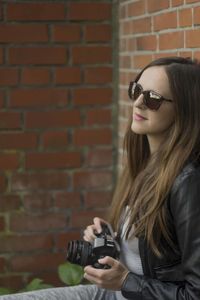 Thoughtful beautiful woman holding camera while leaning on brick wall