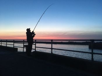 Silhouette man fishing in sea against sunset sky