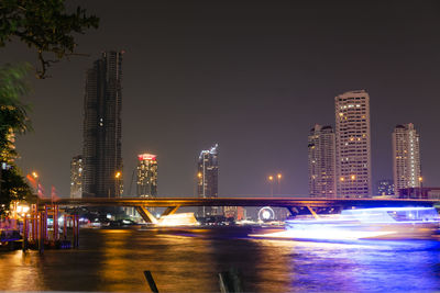 Illuminated modern buildings by river against sky at night