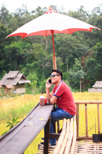 Man talking on phone while having drink at table against trees