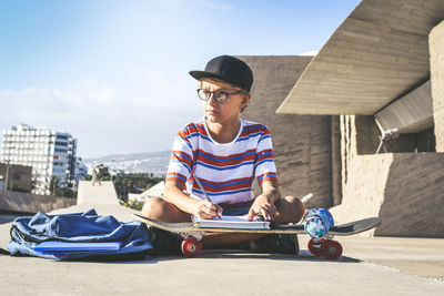 Full length of boy writing in book over skateboard while sitting on road