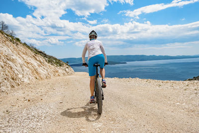 Rear view of man riding bicycle on field by sea against sky