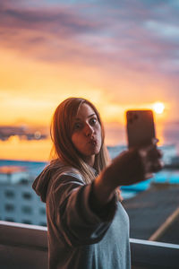 Young woman taking selfie with mobile phone in balcony against sky during sunset