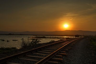 Railroad tracks by lake against sky during sunset