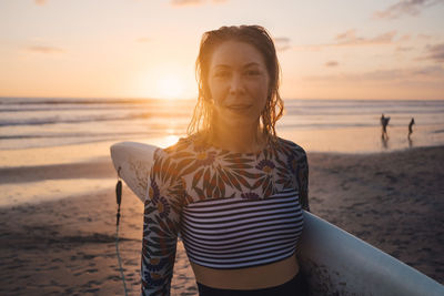 Portrait of mid adult woman with surfboard at beach during sunset