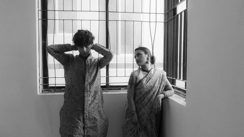 A girl and a boy in traditional wear stand near a window.