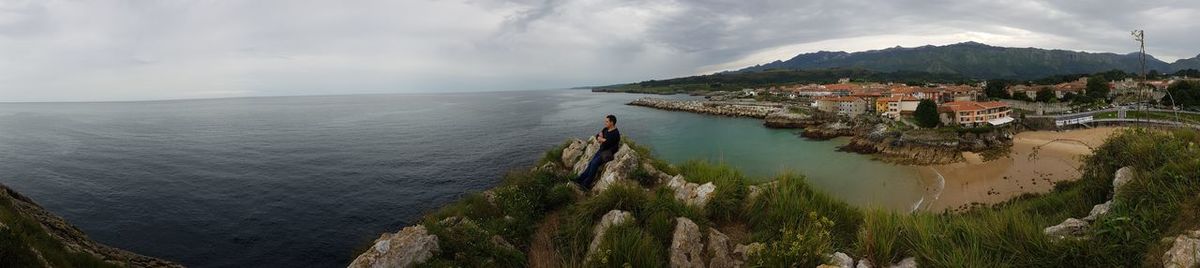 Panoramic view of man sitting on cliff by sea against cloudy sky