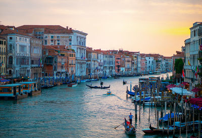 Boats in canal amidst buildings in city against sky during sunset