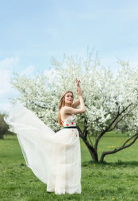 A young blonde in a long white dress poses near a cherry blossom in the garden, a spring landscape.