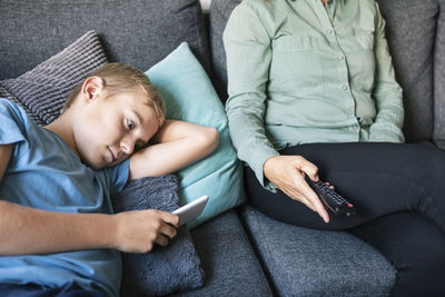 Midsection of woman holding remote control while son using phone on sofa