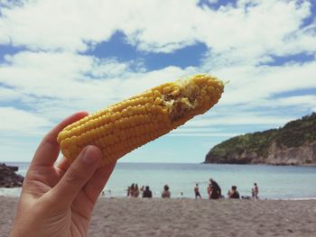 Cropped hand holding corn on the cob at beach against cloudy sky