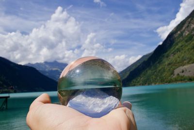 Cropped hand of person holding crystal ball by lake against mountains and sky