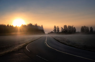 Road by trees against sky during sunset