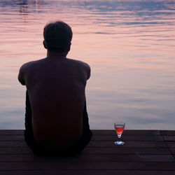 Rear view of shirtless man with drink sitting on pier by lake during sunset