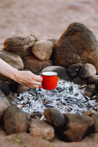 Woman's hand holds a red cup over a campfire in nature. campfire on a hike in the forest. traveling