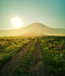 Dirt road amidst field against sky during sunset