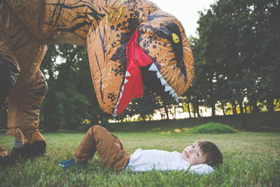 Portrait of boy lying on grassy land while playing with person wearing dinosaur costume in park