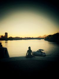 Silhouette man sitting by lake against sky during sunset