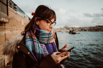 Close-up of woman wearing eyeglasses using smart phone standing by sea