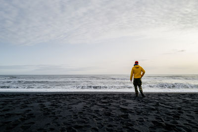 Man in a yellow jacket standing on the black sand beach overlooking the horizon