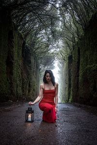 Portrait of mature woman holding lantern while kneeling on road at forest