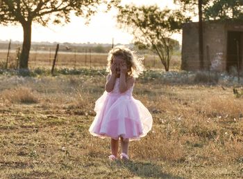 Cute girl in dress covering face with hands while standing on grassy field