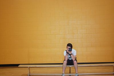 Man photographing through camera while sitting on bench against wall