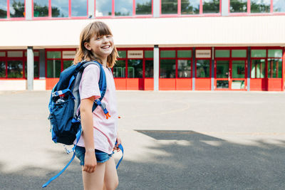A satisfied teenage girl with a school backpack on her back stands in front of the school building