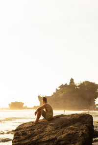 Man sitting on rock by sea against clear sky