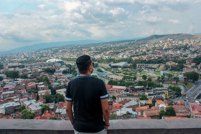 Rear view of mid adult man looking at cityscape while standing by retaining wall against cloudy sky