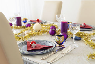 A decorated dining table with burning candles, cutlery, glasses, christmas balls and gold tinsel