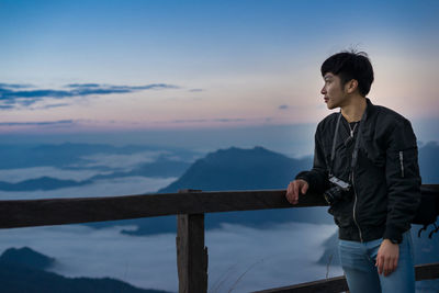 Man standing on railing against sky during sunset