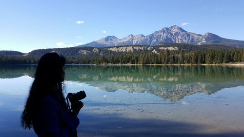 Side view of woman standing with camera at lakeshore against pyramid mountain