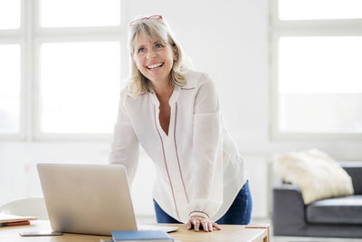 Portrait of happy mature businesswoman with laptop leaning on desk