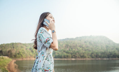 Side view of young woman using smart phone while standing at lakeshore