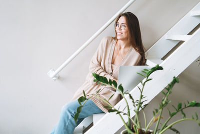 Young woman with long hair in cardigan working on laptop sitting on stairs at home