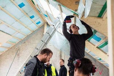 Construction worker drilling roof beam while coworkers standing by ladder at site