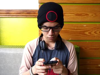 Young man with headphones wearing knit hat using mobile phone while sitting against wall