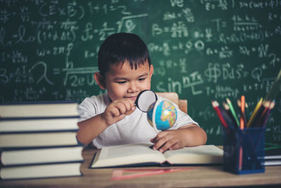 Cute boy studying at table against blackboard