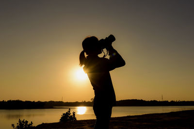 Silhouette man photographing lake against sky during sunset