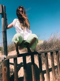 Woman crouching on fence against sky