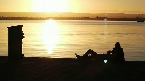 Silhouette woman sitting by lake against sky during sunset