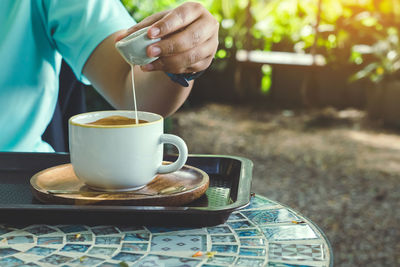 Hand of young woman pouring condensed milk into hot cappuccino on cafe table. 